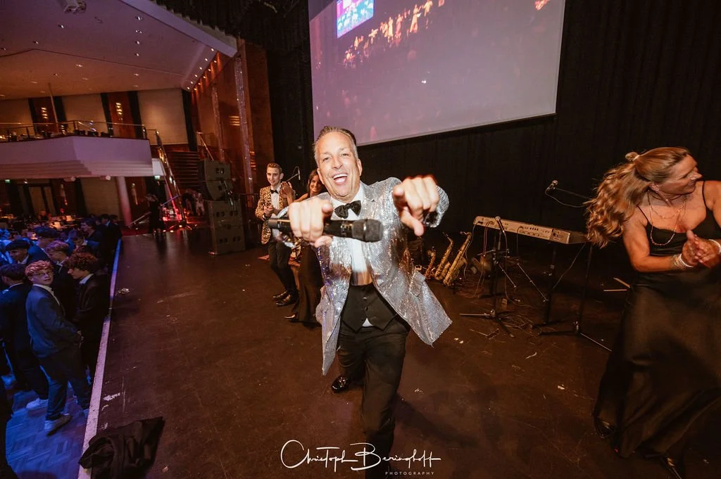 Ein Mann in einem silbernen Jackett hält eine Mikrofon in der Hand auf einer Bühne bei einer Veranstaltung mit einem Live-Musikensemble im Hintergrund.