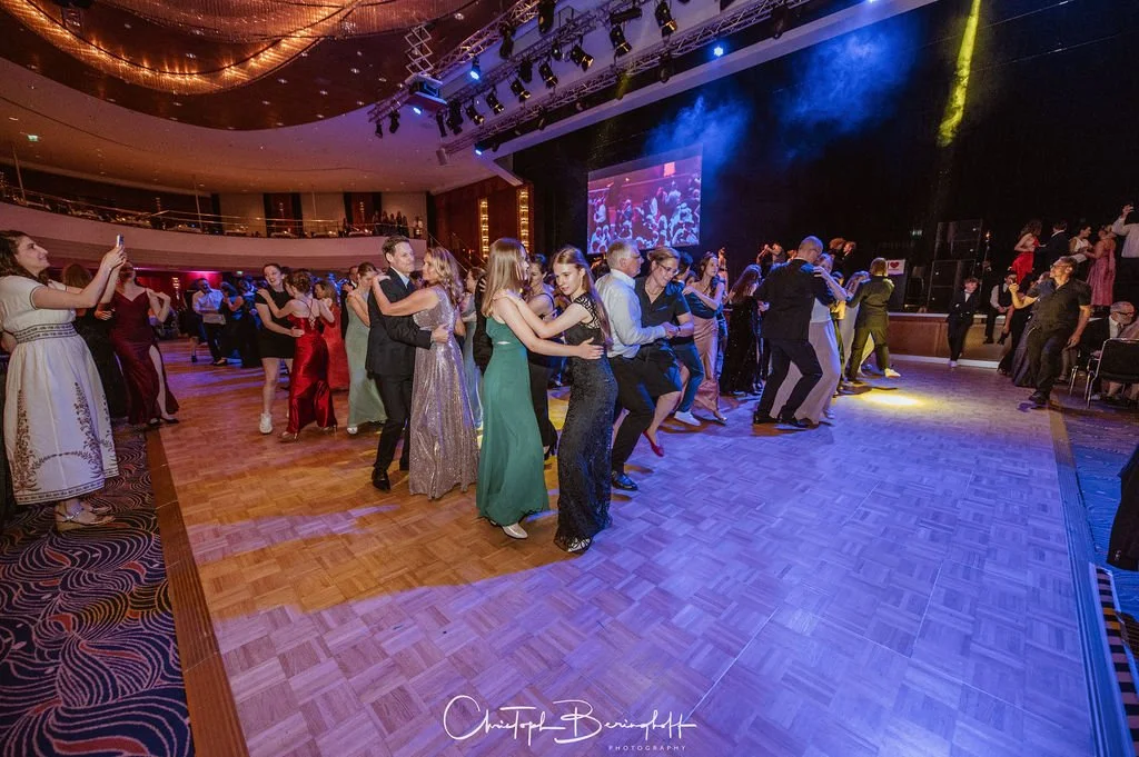 Menschen tanzen auf einer festlichen Party im großen Ballsaal, mit einer Bühne und einem großen Bildschirm im Hintergrund, bunter Beleuchtung und einer eleganten Atmosphäre.