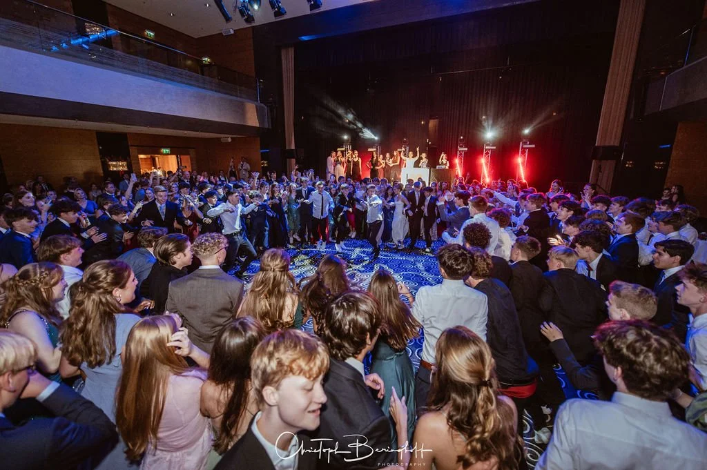 Tanzen auf einer Party in einem großen Veranstaltungsraum mit vielen Gästen, einer Bühne im Hintergrund mit Live-Performance, bunten Lichtern und Disco-Atmosphäre.
