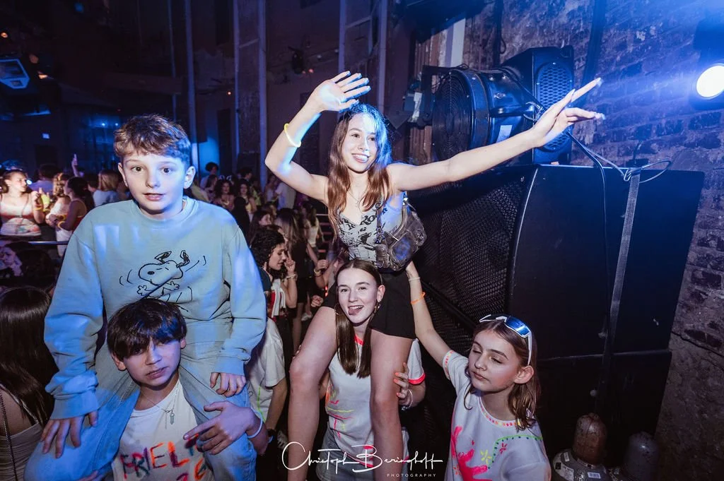 Teenagers are partying at a nightclub; some are dancing while others stand in the background, and a group is posing in front of a speaker.