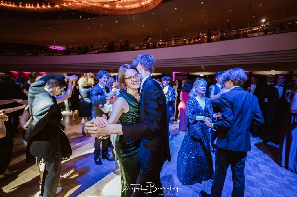 Menschen tanzen auf einer festlichen Party im eleganten Schwarz, Blau und Grün. Der Raum ist stilvoll mit gedämpfter Beleuchtung, und im Hintergrund sind weitere Gäste zu sehen.