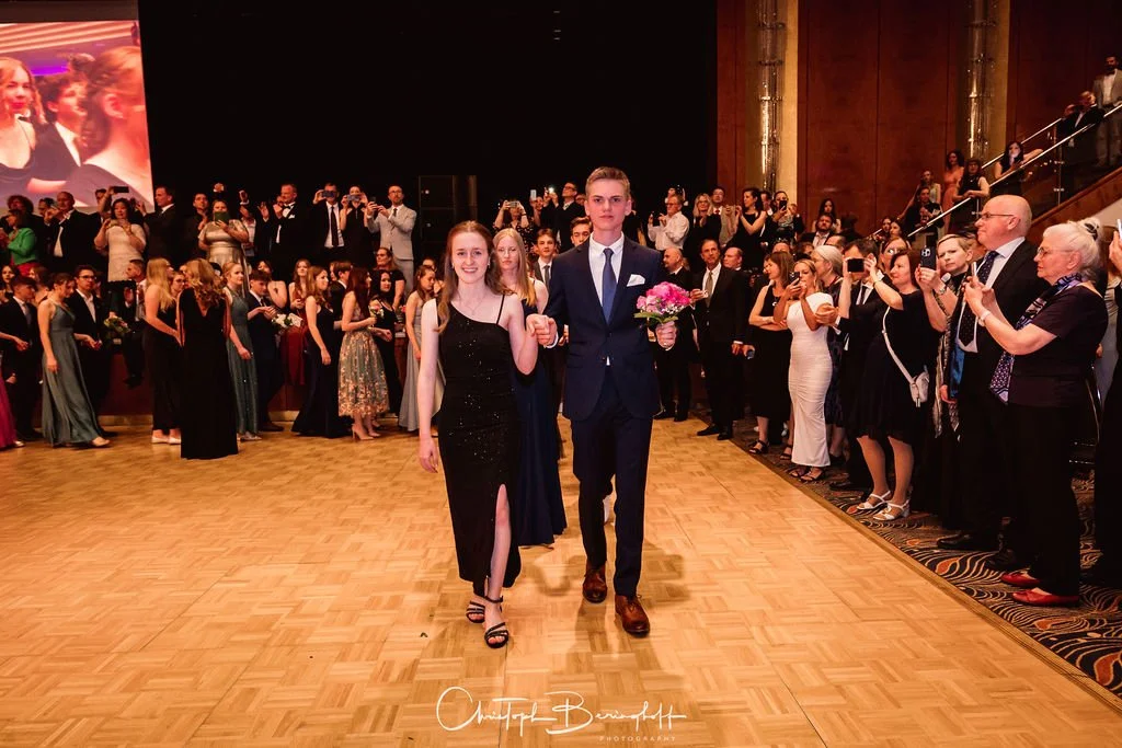 Ein junges Paar bei einer formellen Veranstaltung, die Frau trägt ein schwarzes Kleid mit Spaghettiträgern und einen Schlips, der Mann trägt einen Anzug mit Krawatte und hält einen Blumenstrauß. Viele Menschen im Hintergrund fotografieren und beobach