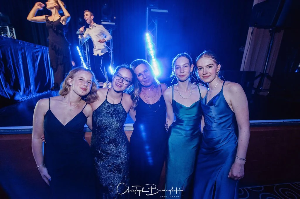 Fünf Frauen in eleganten Abendkleidern posieren vor einer Bühne mit blauer Beleuchtung bei einer Feier.