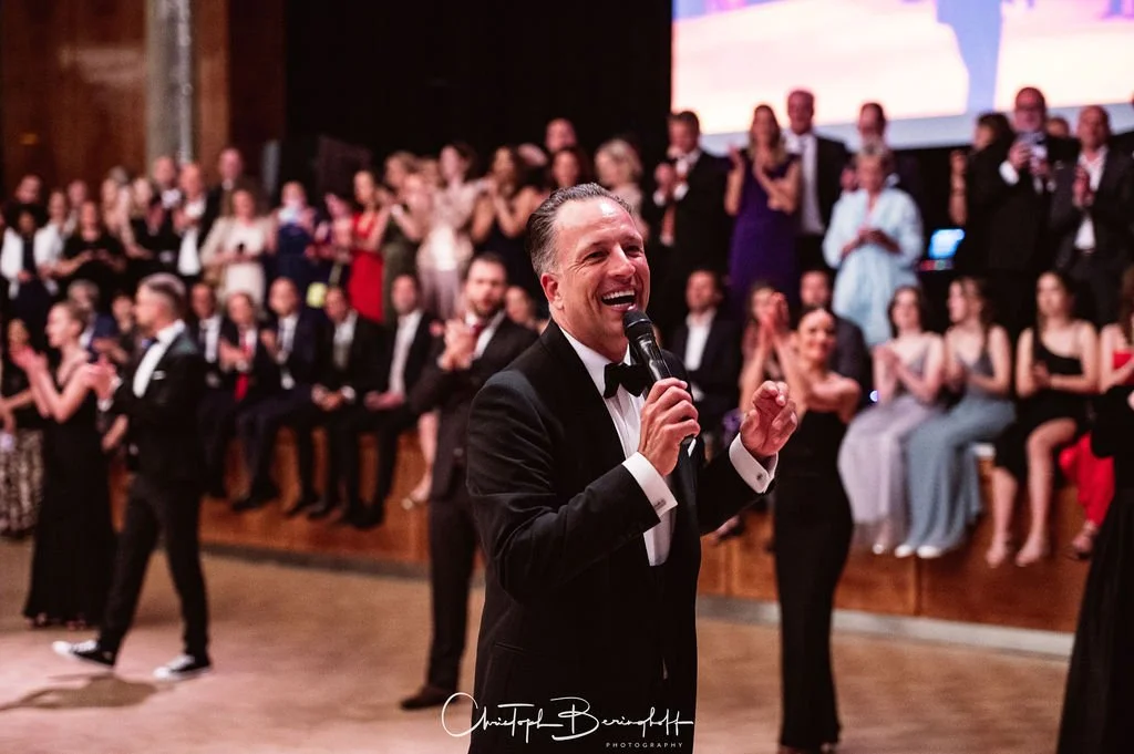 Mann in Anzug und Fliege spricht bei einer Feier ins Mikrofon, im Hintergrund applaudierende Gäste im eleganten Kleid und Anzug, festliche Stimmung.
