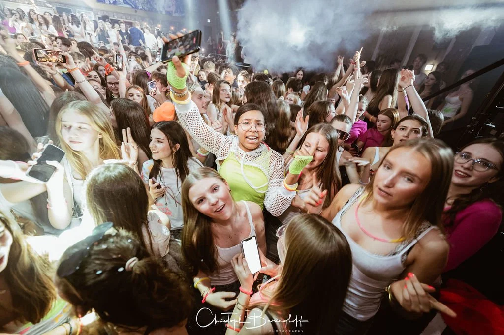 People celebrating at a party; many are taking photos with their cell phones, some are dancing, with colorful lights and smoke in the background.