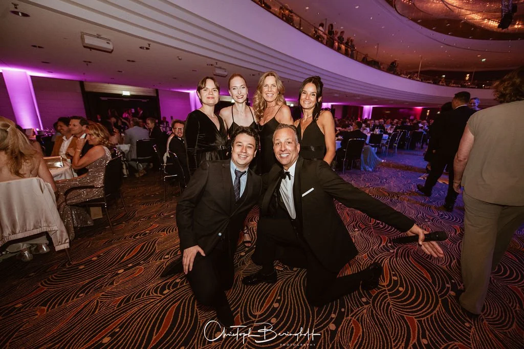 Menschen bei einer festlichen Veranstaltung in einem Ballsaal, vier Frauen im schwarzen Kleid und zwei Männer in Anzügen posieren für ein Foto, im Hintergrund sind weitere Gäste und der elegante Veranstaltungsraum zu sehen.
