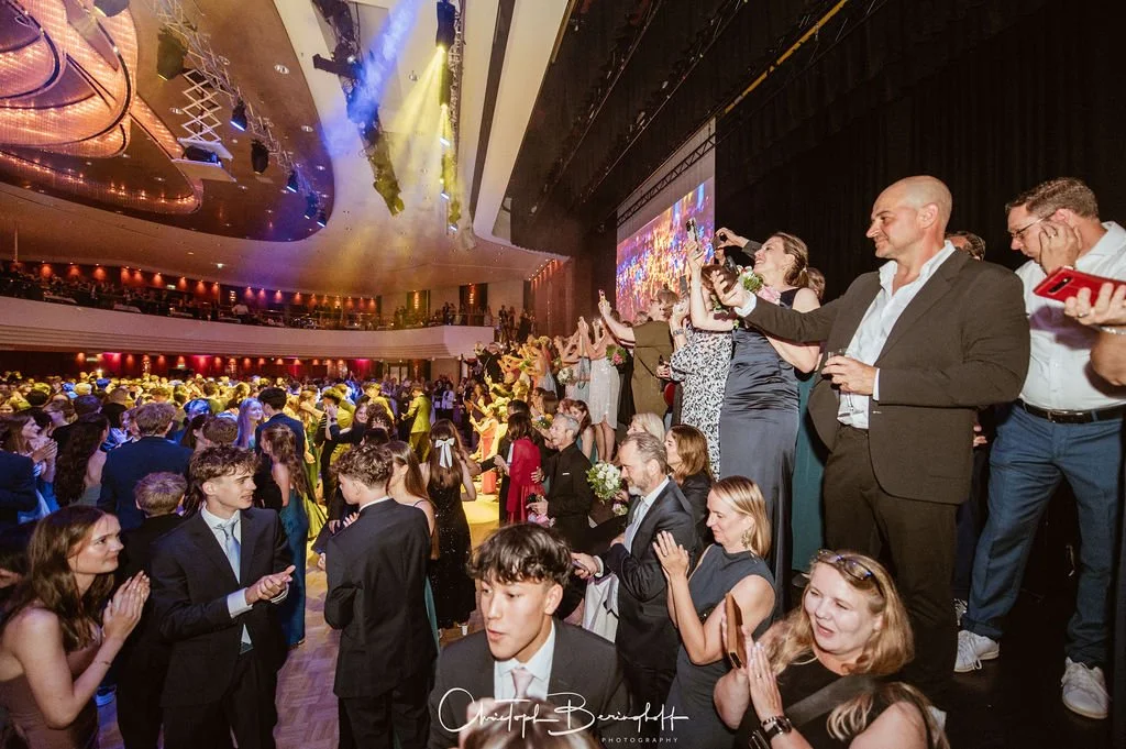 Menschen feiern auf einer Tanzfläche bei einer festlichen Veranstaltung im großen Saal, einige nehmen Fotos auf, Glamour und Bewegung, festliche Kleidung, bunte Beleuchtung.