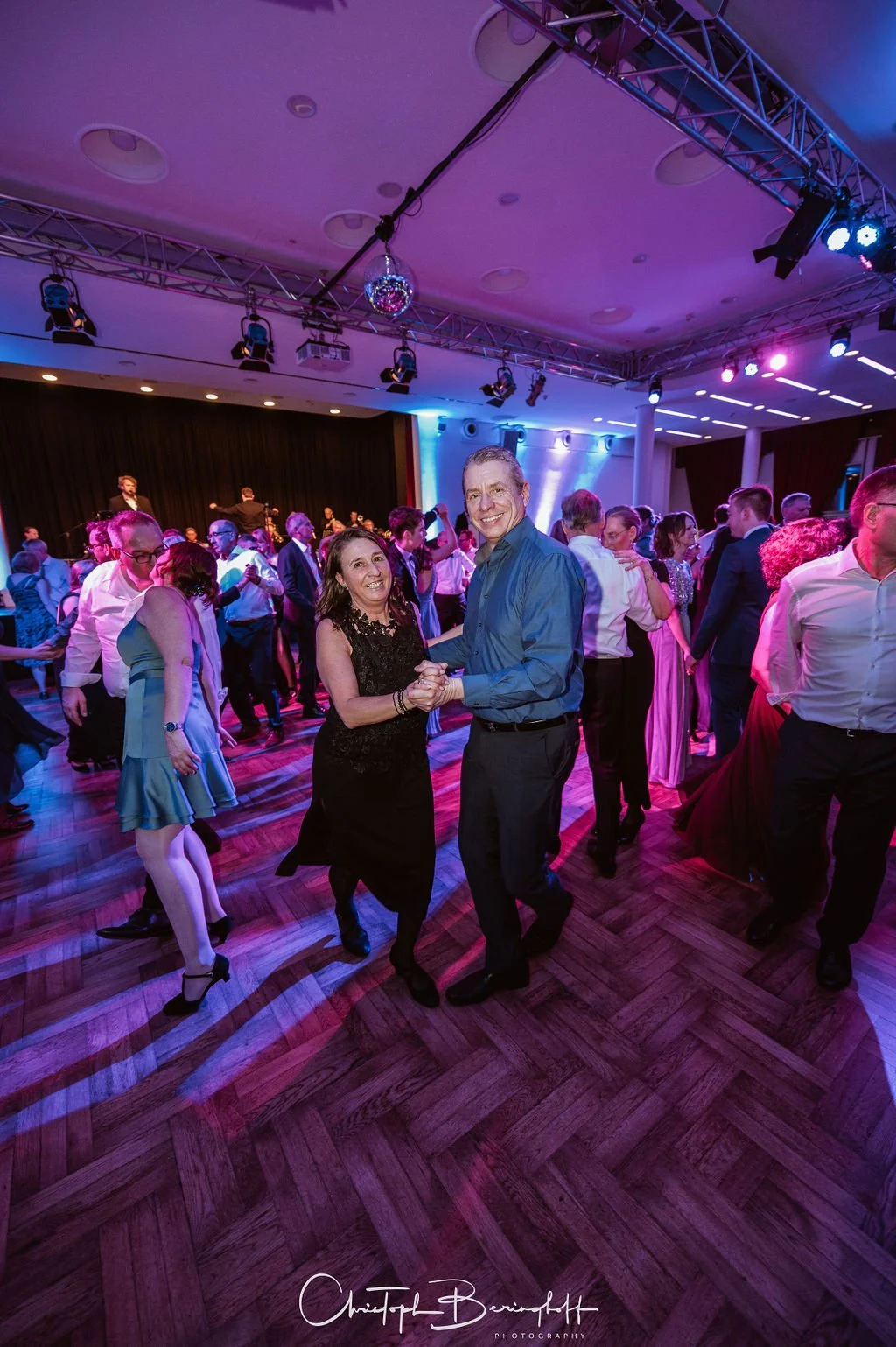 Menschen tanzen bei einer Party unter buntem DJ-Licht, im Vordergrund ein Tanzpaar, im Hintergrund eine Musikgruppe auf der Bühne.