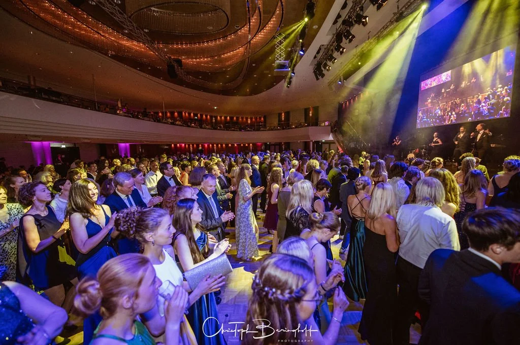 Menschen auf einem Gala-Abend in einem großen Konzertsaal bei einer live-Performance. Die Gäste stehen, einige klatschen, während auf der Bühne Musiker auftreten. Das Ambiente ist elegant, mit bunten Lichtern und einem großen Bildschirm.