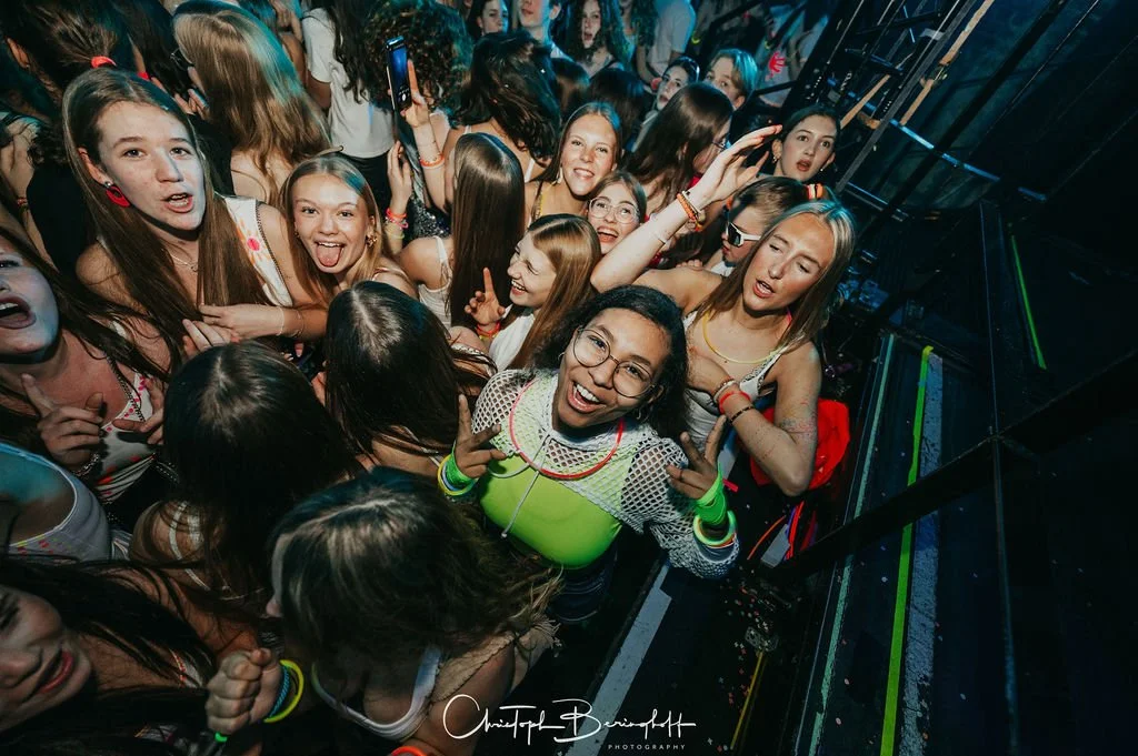 A group of young people are dancing at a party; many are wearing neon accessories and smiling at the camera.