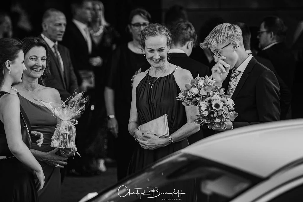 Mehrere Menschen, darunter zwei Frauen und ein Mann, bei einer formellen Veranstaltung. Eine Frau hält einen Blumenstrauß, ein Mann trägt einen Blumenstrauß, und alle scheinen sich zu unterhalten.