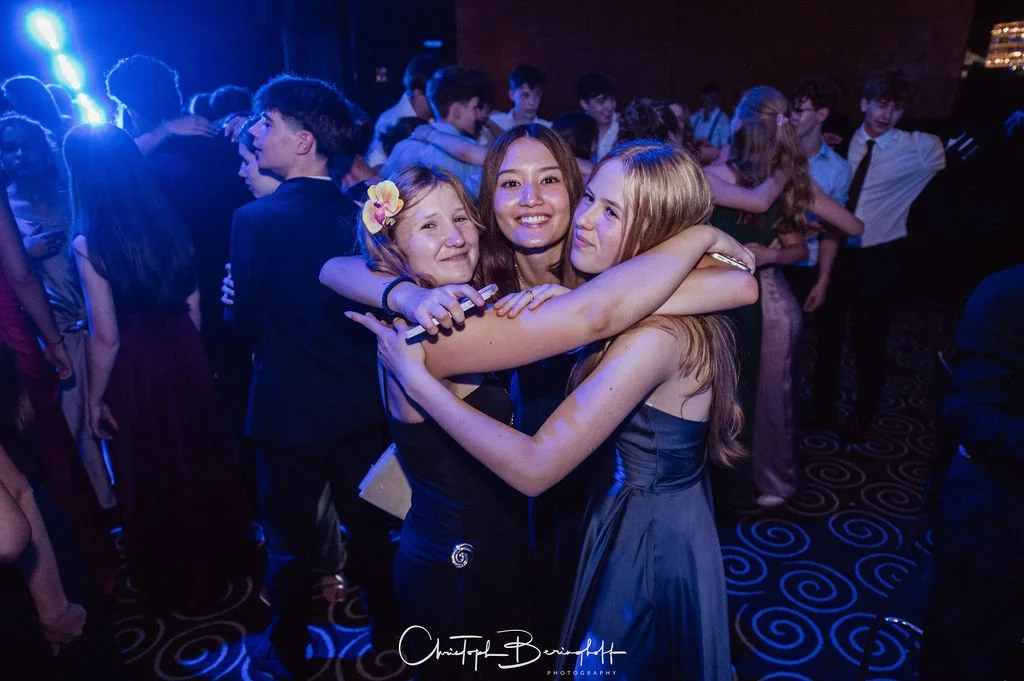 Grupp of teenage girls hugging at a dance party, smiling with purple and blue lighting, in formal dresses.