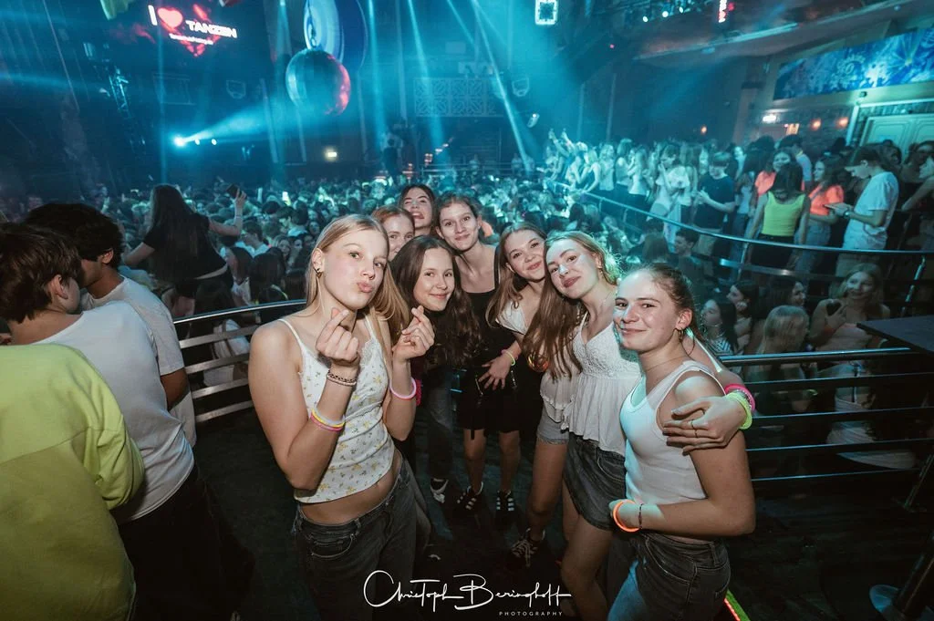 A group of young women poses in a nightclub with a dance floor and colorful lights in the background.