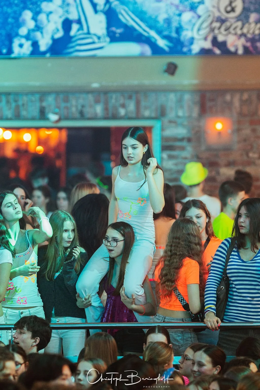 A group of young people in a dark room; some are sitting, others are standing; one woman is sitting on another person’s shoulders; everyone is dressed casually; in the background is a bar with warm lighting.