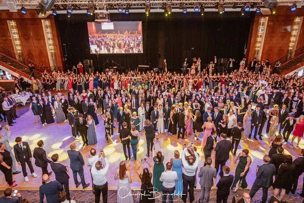 Festliche Tanzveranstaltung in einem großen Saal mit vielen Gästen, die im formellen Outfit tanzen und Fotos machen, mit einer Bühne im Hintergrund.