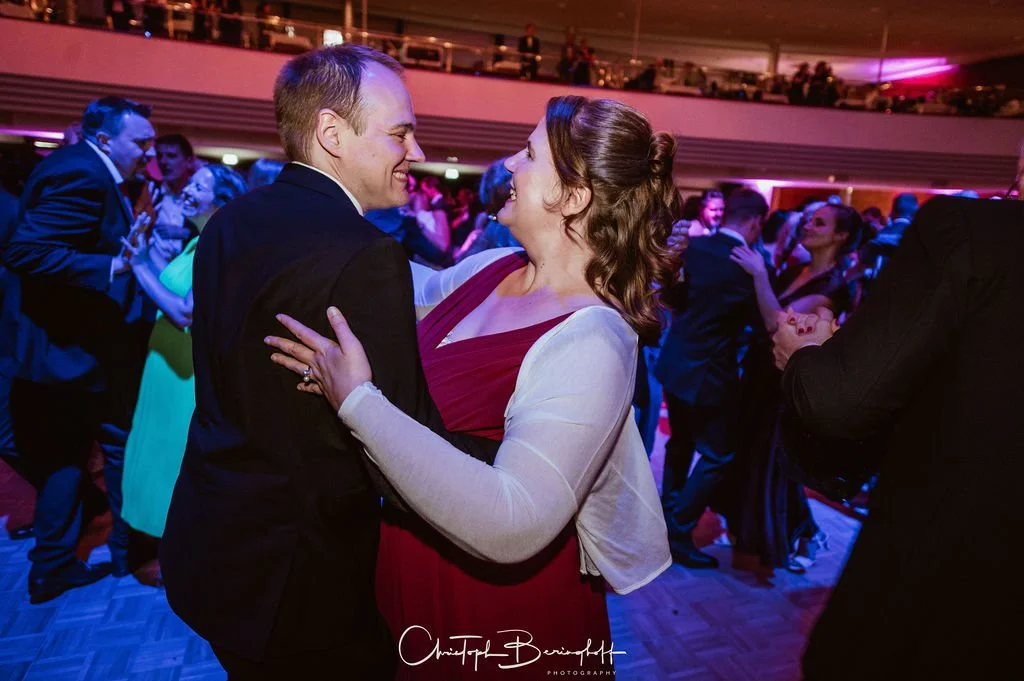 Ein Mann und eine Frau tanzen eng auf einer Party im abendlichen Rahmen, umgeben von anderen tanzenden Gästen.