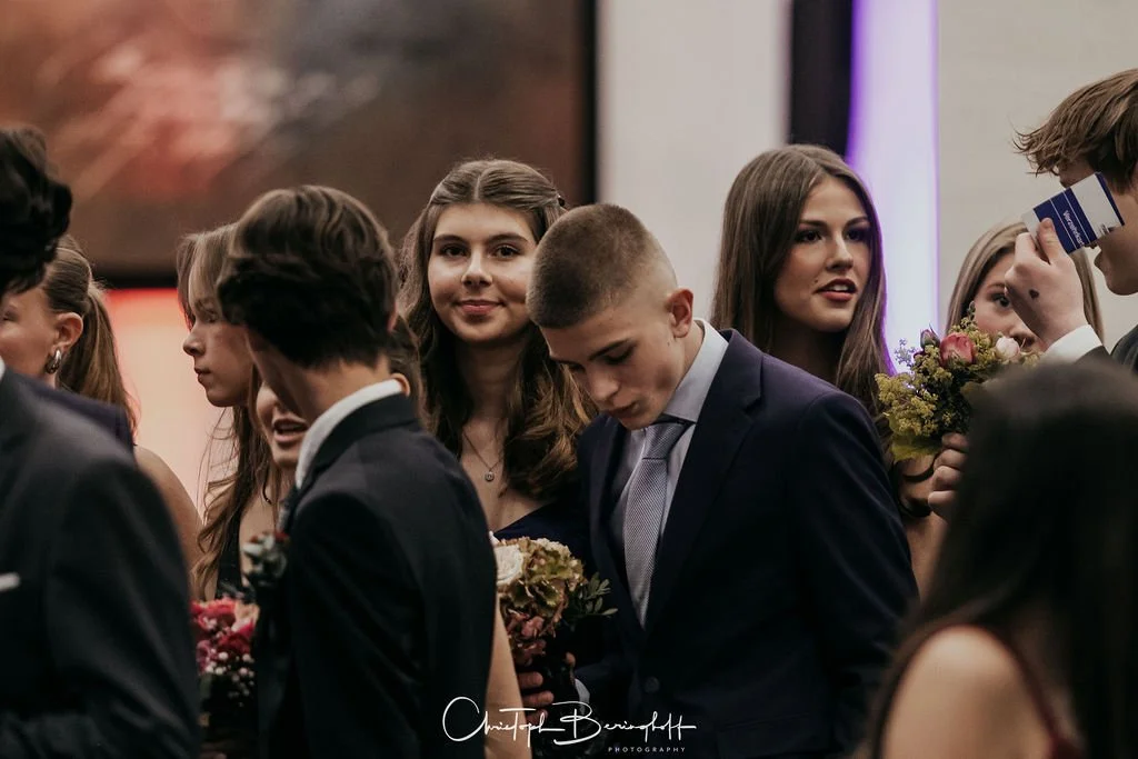 Junge Menschen in formeller Kleidung bei einer Veranstaltung, einige halten Blumen, im Hintergrund ein Bildschirm und Lichter.