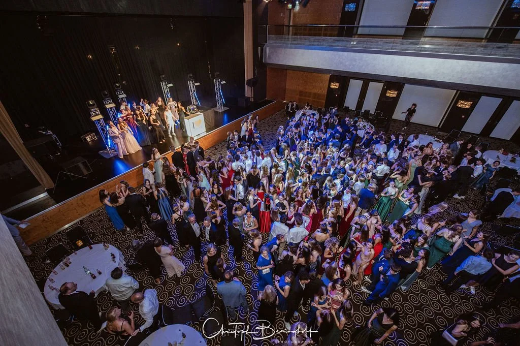 Tanzveranstaltung mit vielen tanzenden Paaren auf der Tanzfläche, auf einer Bühne eine Gruppe von Menschen, die auf einer Party oder Gala auftreten, im Raum sind Tische mit Gästen, dunkler Teppich mit spiralförmigem Muster, modern eingerichteter Vera
