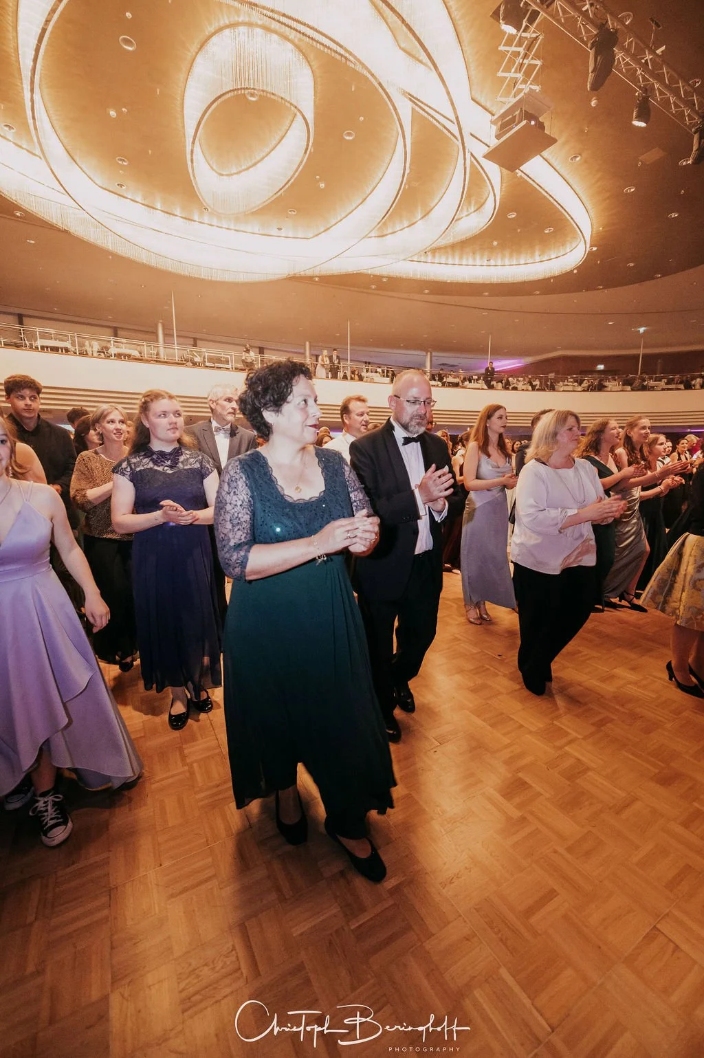 Menschen tanzen bei einer formellen Veranstaltung in einem eleganten Saal mit moderner Beleuchtung und Holzparkett.
