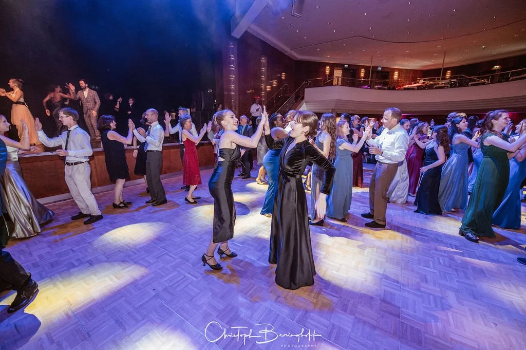 Menschen tanzen in eleganten Kleidern und Anzügen auf einem Tanzparkett in einem festlichen Ballsaal