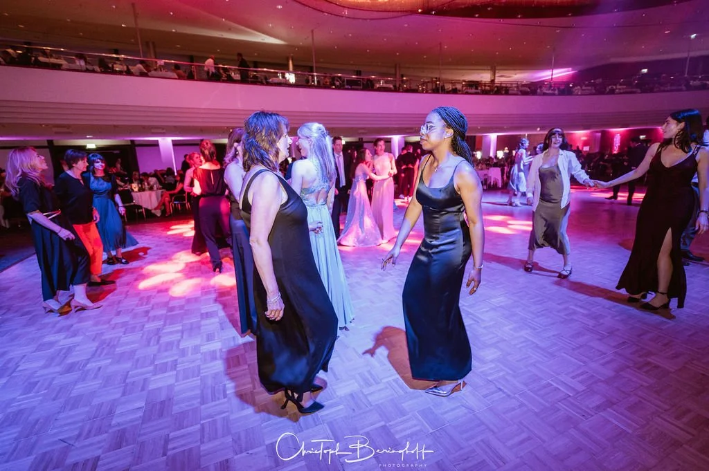Menschen tanzen auf einer Party oder Gala in einem elegant dekorierten Saal mit bunten Lichteffekten.