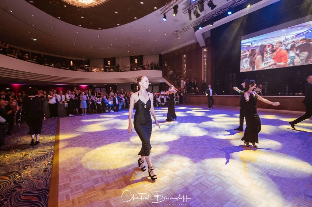Tanzveranstaltung in einem Ballsaal mit Leuten, die tanzen und zuschauen, große Leinwand im Hintergrund, bunte Lichter und eine elegante Atmosphäre.