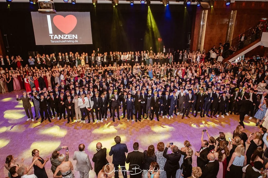 Große Gruppe junger Menschen in formeller Kleidung auf Tanzveranstaltung in einem Saal, mit Tanzschule Freiling.de Logo im Hintergrund.