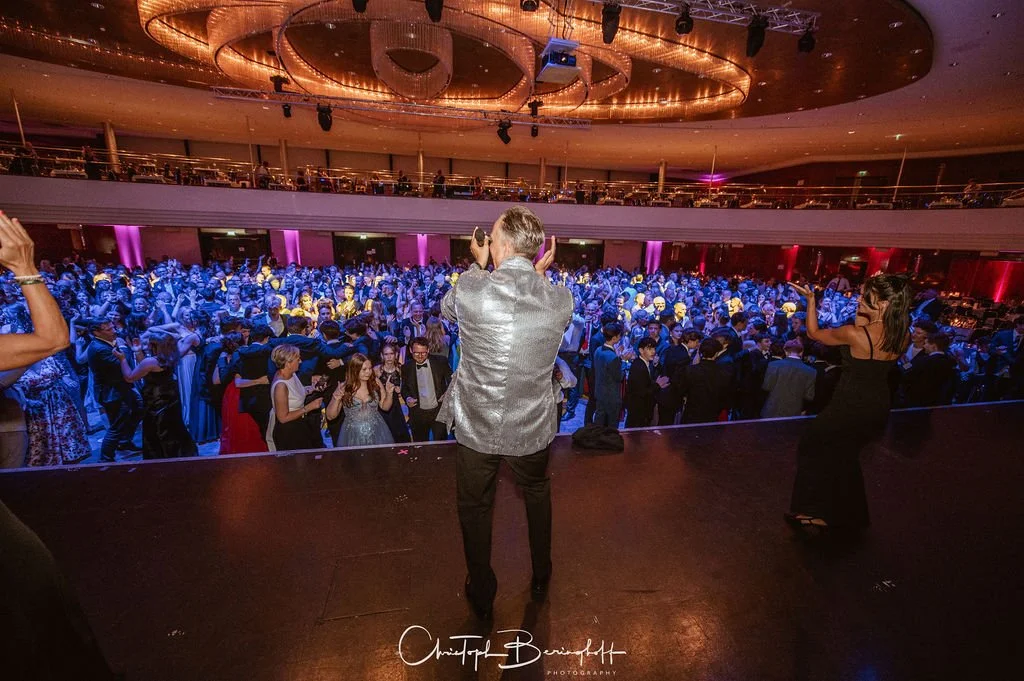 Bühne mit einem Jungen in einem silbernen Jacke und einem Mädchen in einem schwarzen Kleid, die vor einer großen tanzenden Menschenmenge in einem Veranstaltungssaal stehen.
