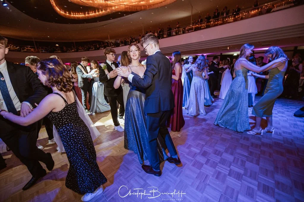 Menschen tanzen bei einer formellen Veranstaltung im eleganten Ballsaal, viele tragen Abendkleider und Anzüge.