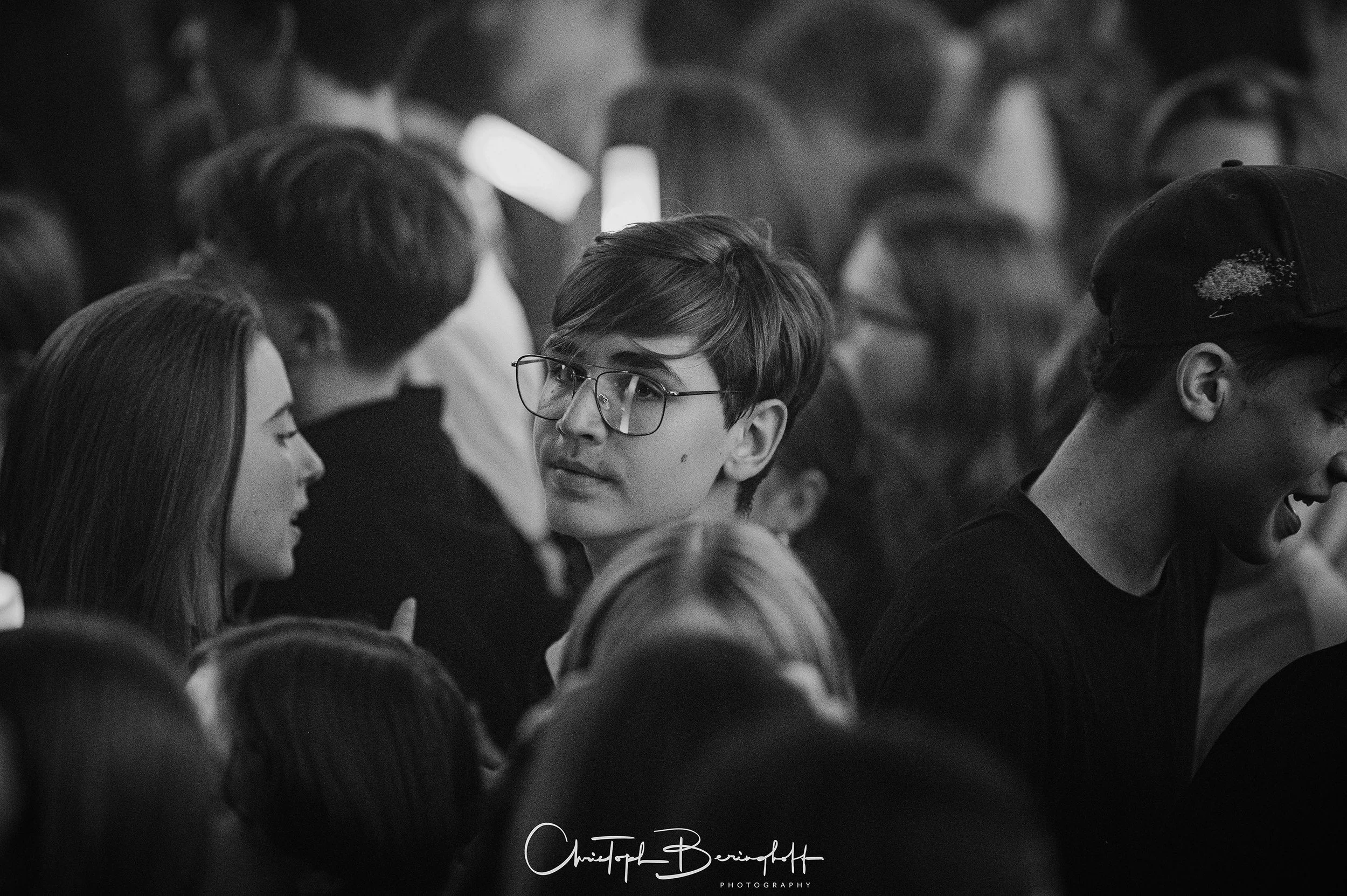 Schwarz-weiß-Foto einer Menschenmenge, im Mittelpunkt ein junger Mann mit Brille, der eine Unterhaltung mit einer jungen Frau hat, umgeben von weiteren Personen, die in einem Raum stehen.