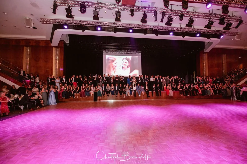 Großer Ballsaal mit vielen elegant gekleideten Menschen, die auf einer Bühne im Hintergrund sitzen, an einem formellen Ereignis oder Gala. Es gibt einen großen Bildschirm, der ein Bild von zwei lachenden Frauen zeigt, und die Tanzfläche im Vordergrun