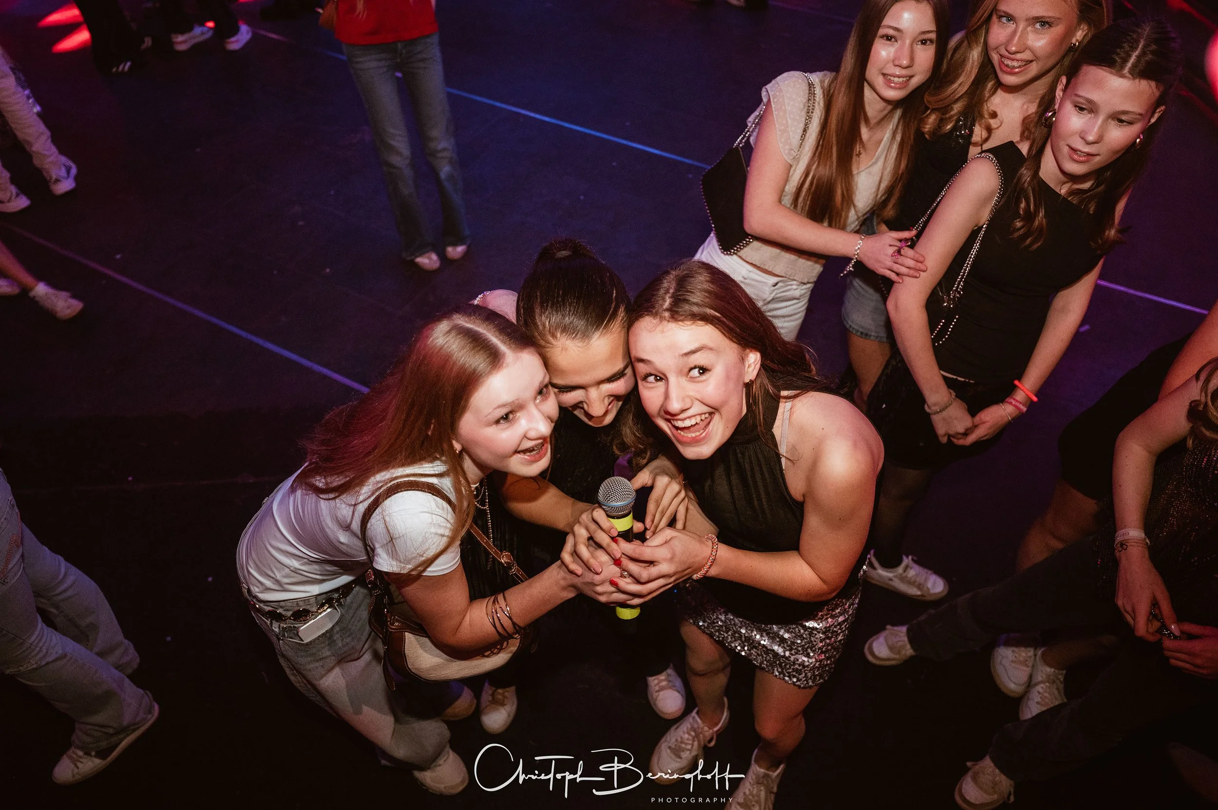 Gruppe von jungen Frauen, die glücklich lachen und zusammenhalten, während sie auf einer Party singen oder sprechen, mit einem Mikrofon in der Hand, umgeben von anderen Partygästen, in einem dunklen Raum mit bunten Lichtern.