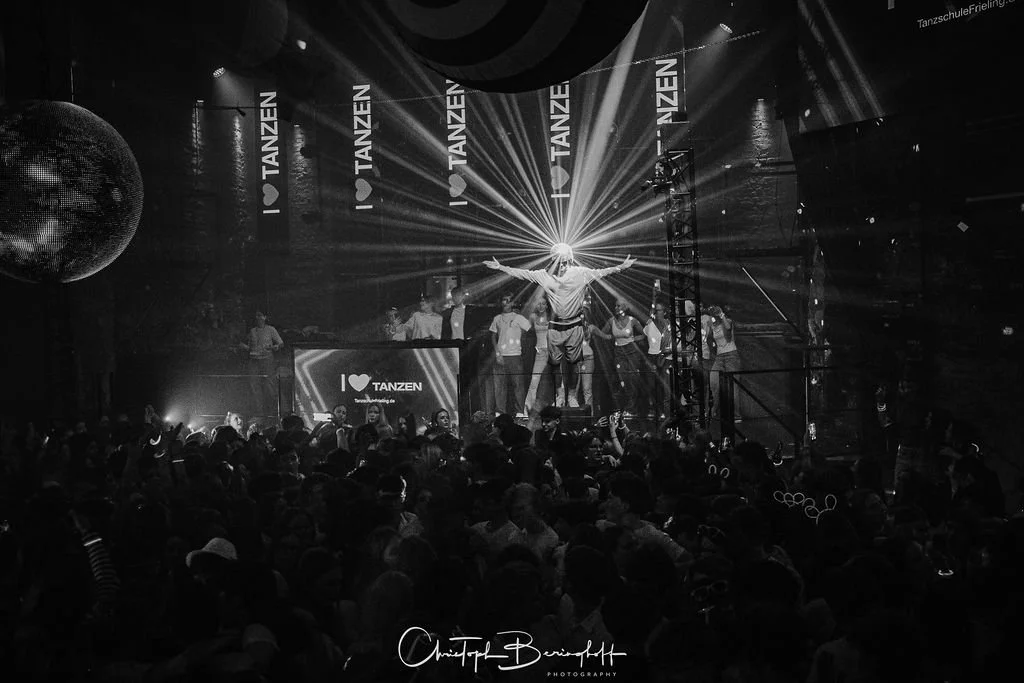 A dance party with a DJ on set, surrounded by people dancing, a stage with lighting and a large disco ball, in black and white.