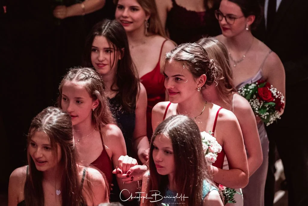 Gruppe von jungen Frauen bei einer Feier, einige halten Blumen, alle tragen elegante Kleidung.