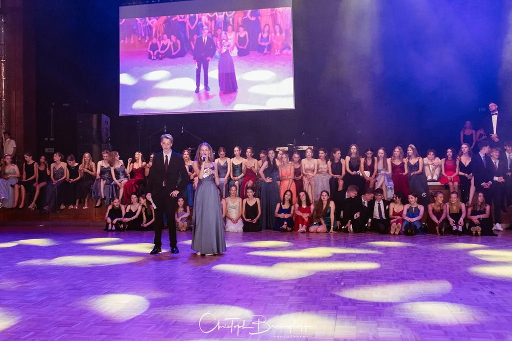 Junge Männer und Frauen bei einer formellen Tanzveranstaltung oder Ball, sitzend und stehend in einem großen Saal, mit einem großen Bildschirm im Hintergrund, der eine ähnliche Szene zeigt.