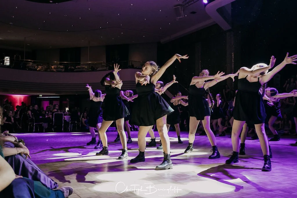 Tänzerinnen in schwarz kleidern Performing auf einer Bühne mit Pink- und Schwarzlicht, Publikum im Hintergrund.