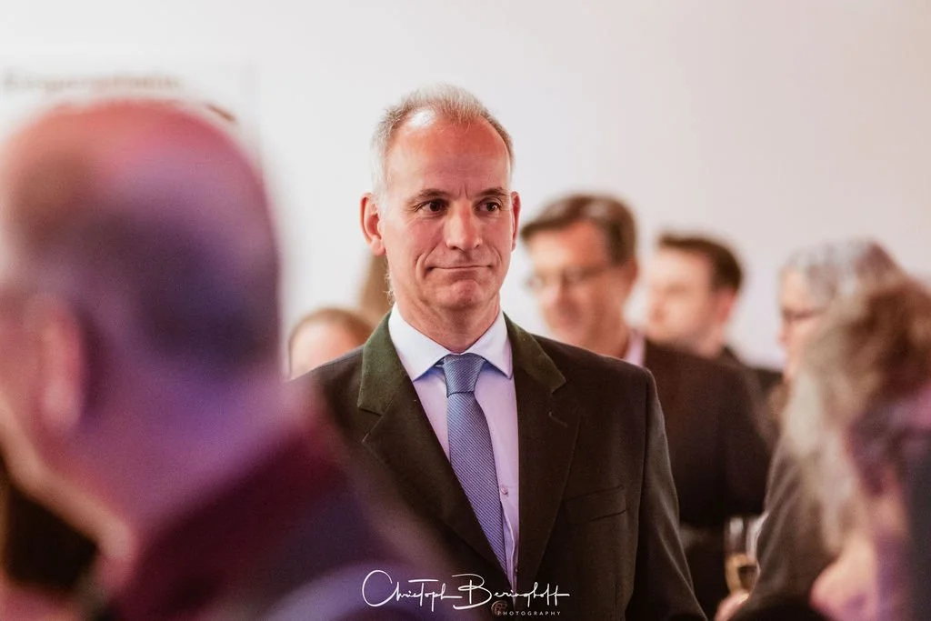 Ein Mann in einem schwarzen Anzug und blauer Krawatte steht bei einer formellen Veranstaltung, umgeben von weiteren Menschen, im Hintergrund unscharf.