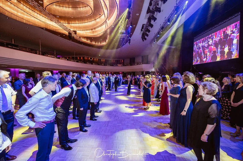 Menschen bei einer formellen Veranstaltung im großen Saal, viele stehen in Reihen, die auf die Bühne blicken, mit einer großen Leinwand im Hintergrund, bunte Lichter und eine elegante Atmosphäre.