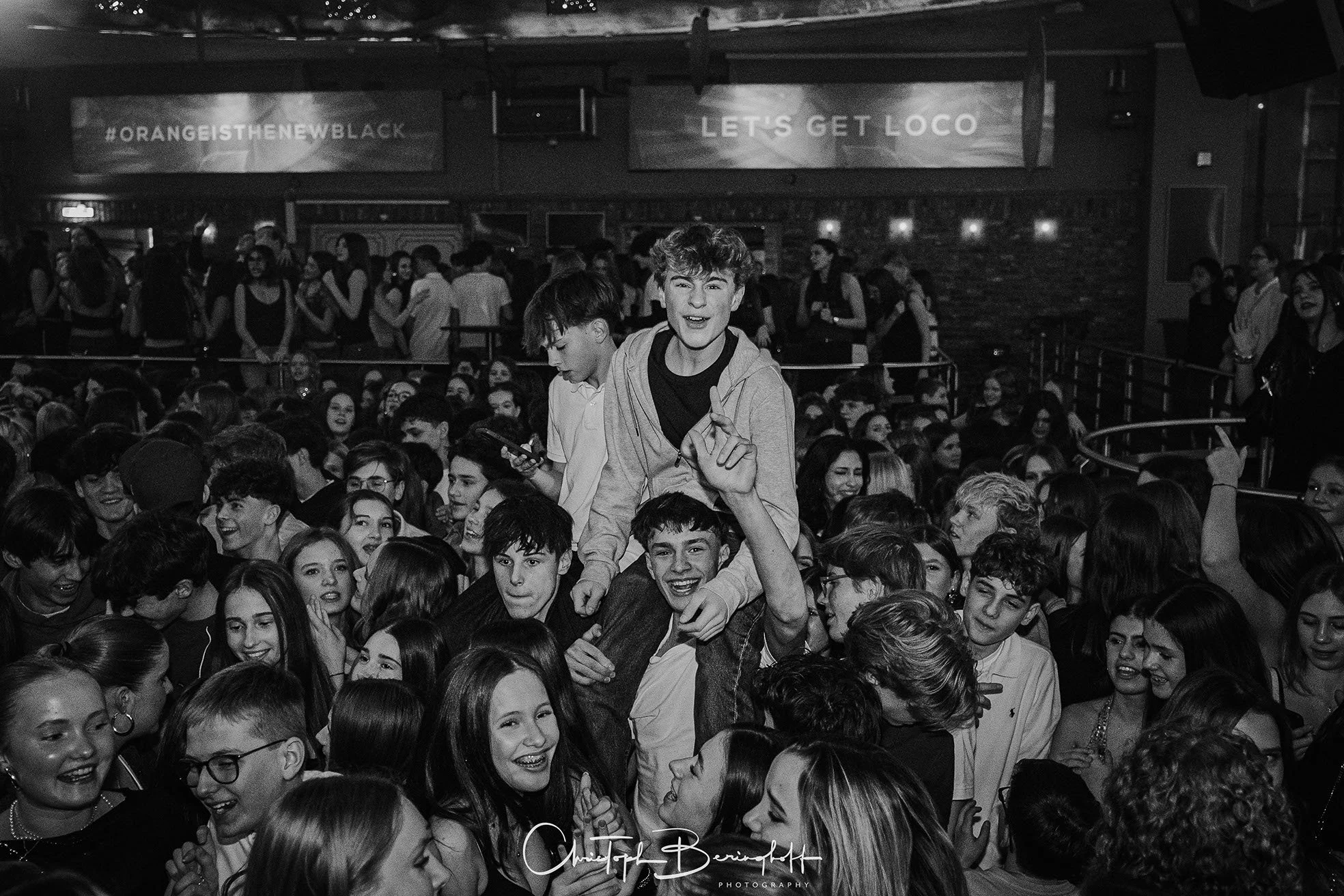 Eine Menschenmenge auf einer Party, bei der einige Jugendliche auf den Schultern anderer sind, in einem dunklen Raum, schwarz-weiß Foto.