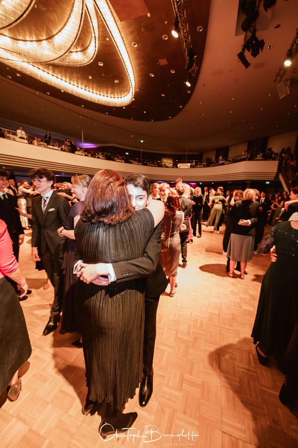 Ein Tanzball mit vielen Gästen in einem großen Saal, mindestens eine Person umarmt eine andere beim Tanzen, der Raum ist festlich dekoriert und gut beleuchtet.