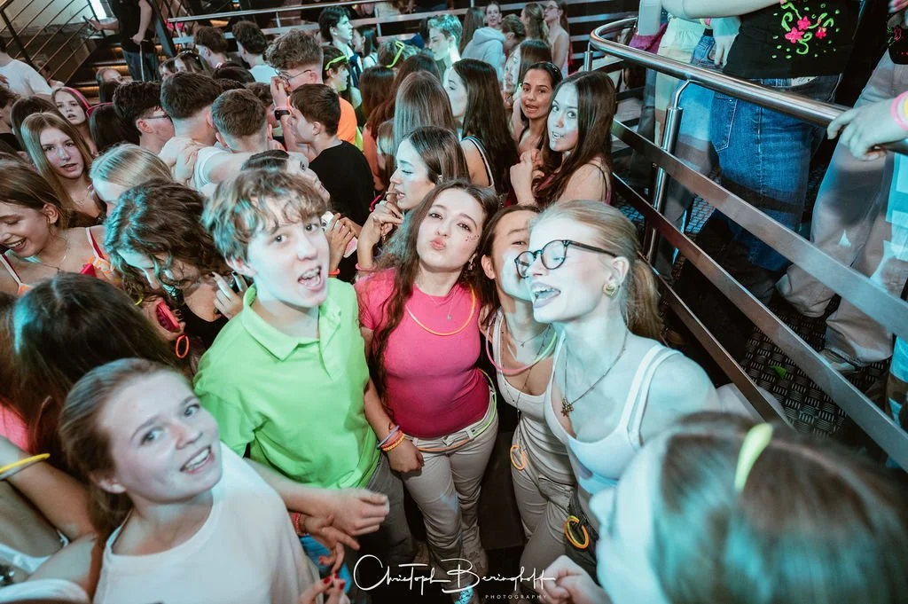 Young people standing together at a party or event, laughing and having fun, with a crowd of people in the background.