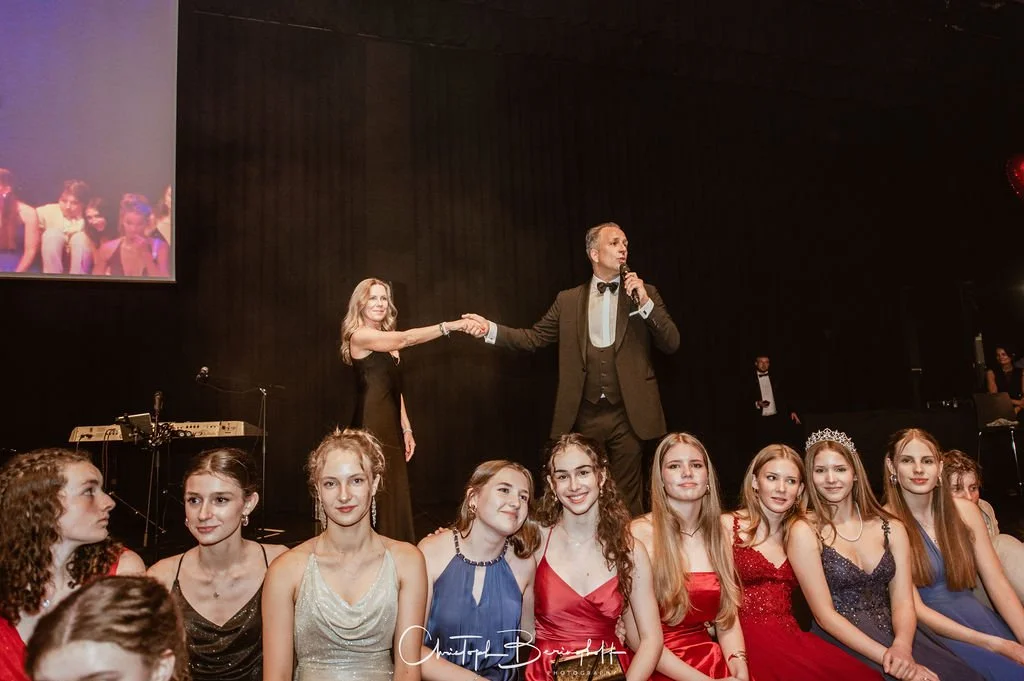 Jugendliche bei einer Schönheitswettbewerb-Veranstaltung, einige tragen Kronen und elegante Kleider, auf der Bühne im Hintergrund ein Moderator in Anzug, Menschen im Publikum
