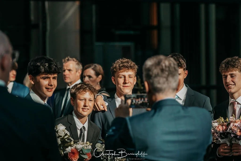 Jugendliche und Männer in formeller Kleidung posieren für ein Foto bei einer Veranstaltung, einige halten Blumen, im Hintergrund eine moderne Gebäude-Architektur.