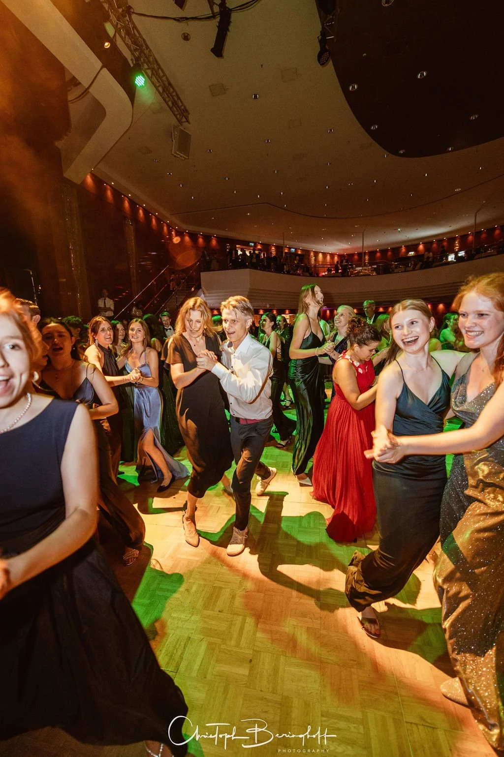 Menschen tanzen auf einer festlichen Veranstaltung in einem großen Saal mit Tanzfläche, einige lachen und haben Spaß, die Atmosphäre ist lebhaft und fröhlich.