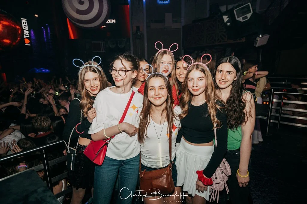 Five women and two girls wearing glowing bunny-ear accessories are partying in front of a DJ booth with illuminated stages. Many other party guests can be seen in the background.