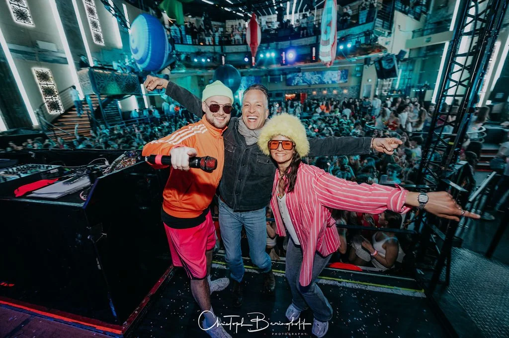 Three people are posing on a stage during a party at a club, with a large crowd in the background.