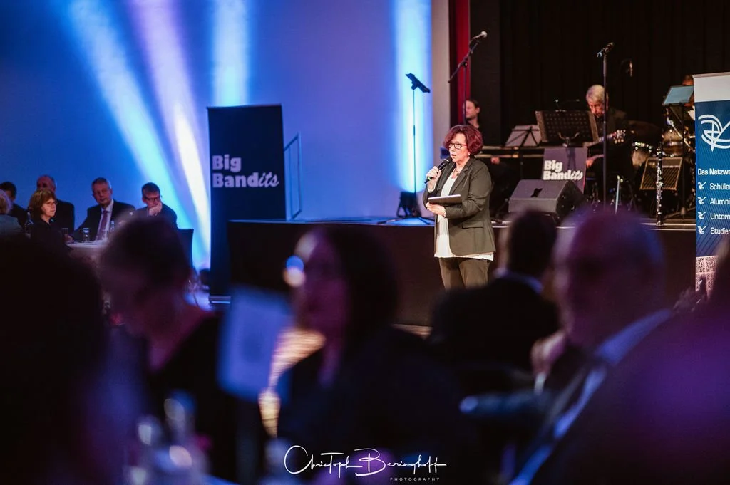 Eine Frau spricht bei einer Veranstaltung auf einer Bühne, im Hintergrund eine Band mit Musikinstrumenten, im Publikum sitzen Menschen im Saal.