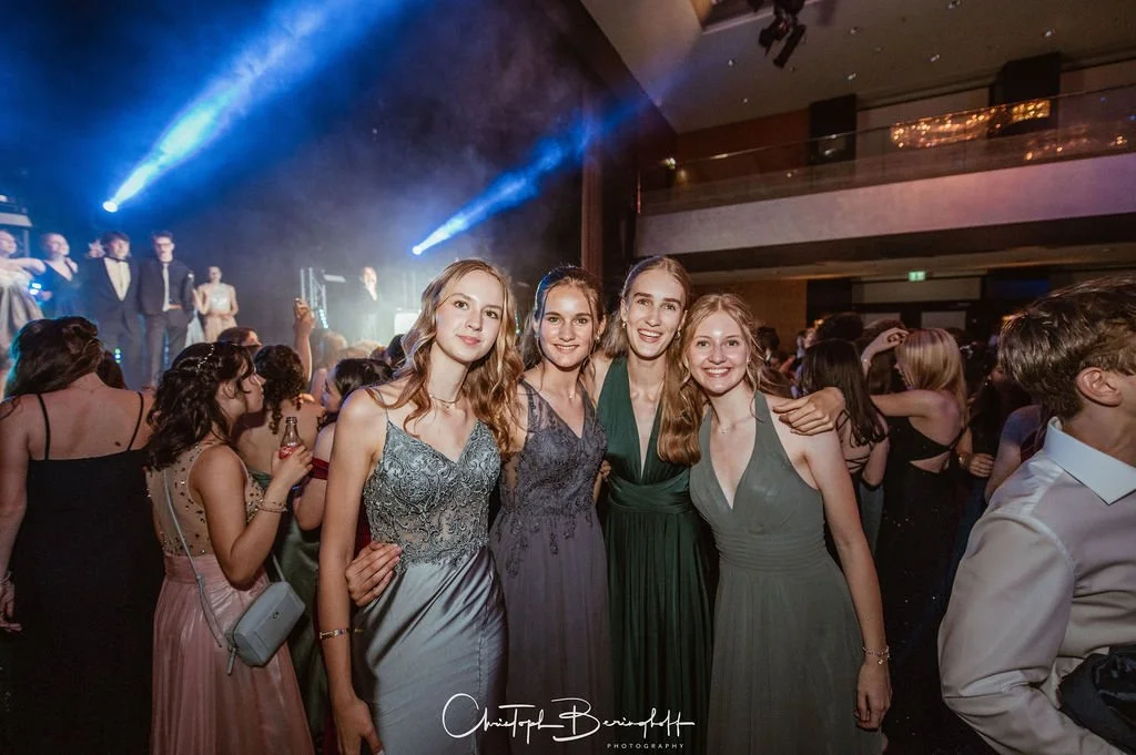 Vier junge Frauen in eleganten Kleidern, die bei einer Party oder Gala tanzen und Fotos machen, mit einer Bühne im Hintergrund, auf der Sänger oder Performer stehen.