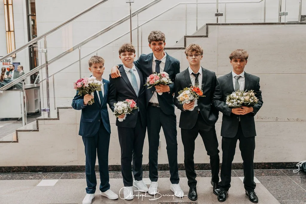 Fünf Jungen in Anzügen, die Blumen halten, stehen in einem Innenraum vor einer Treppe.