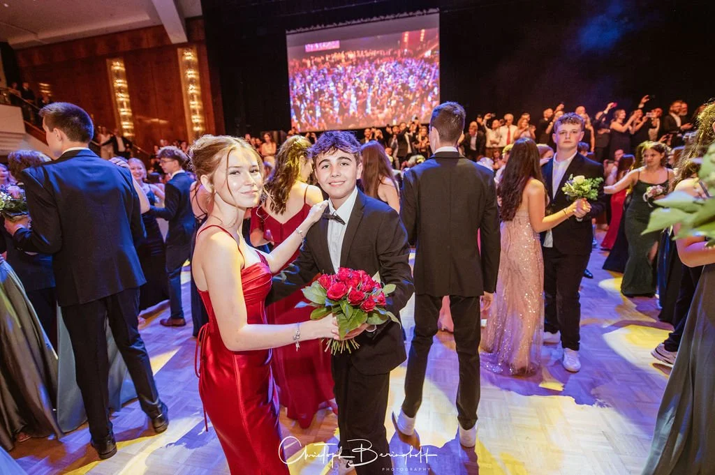 Junge Frau in rotem Abendkleid und Junge Mann in dunklem Anzug mit Blumenstrauß auf Tanzveranstaltung im Saal
